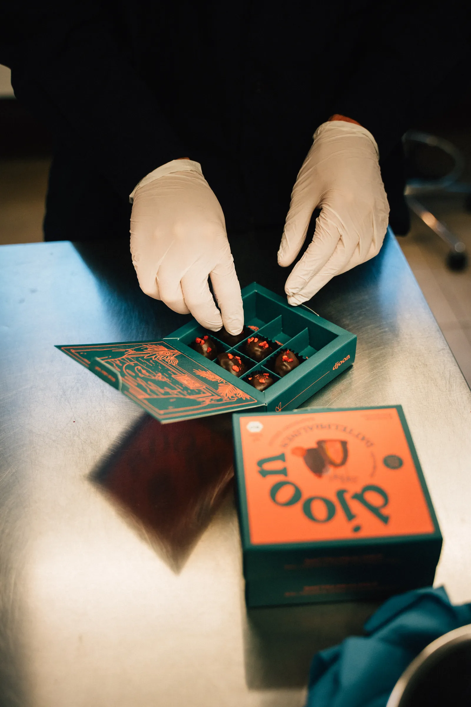 Hände in Handschuhen packen Pralinen in eine orange-grüne Schachtel von djoon.